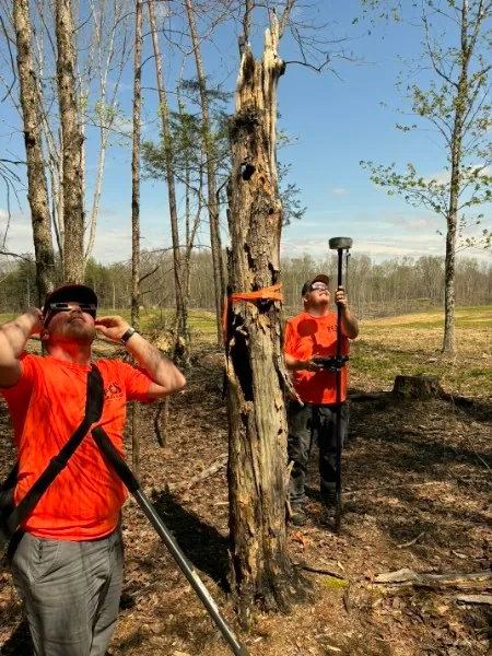 Two Doss Land Surveying field crew members working together at a GPS rover in wooded terrain, one wearing eclipse glasses during the 2024 solar eclipse.
