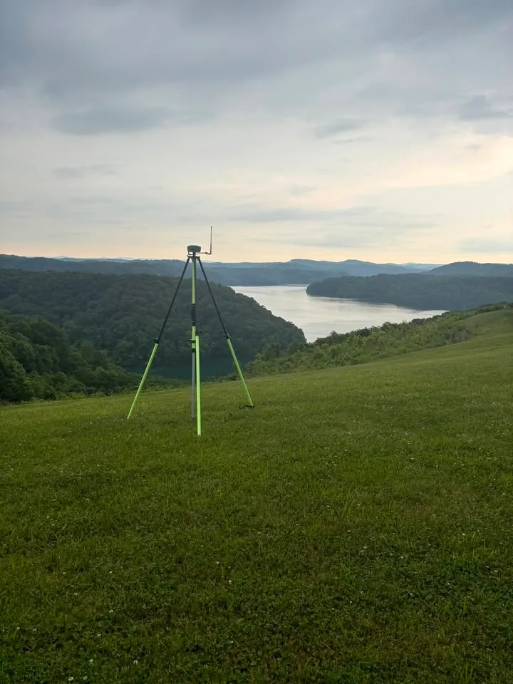 A Doss Land Surveying GPS base receiver set up on a tripod in an open pasture, overlooking a river valley in Middle Tennessee at first light.